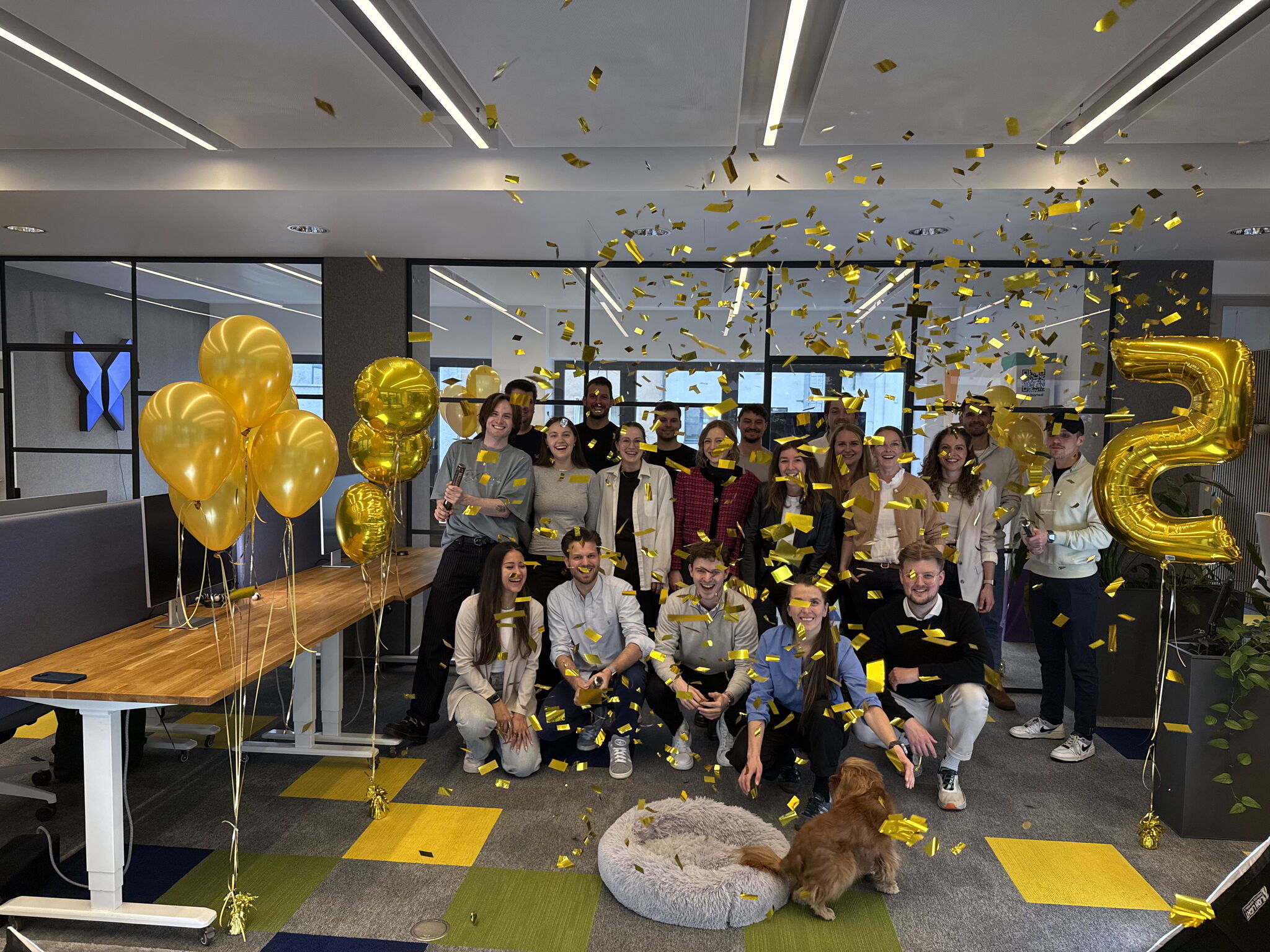 Team celebrating a milestone in a modern office with gold balloons and confetti.