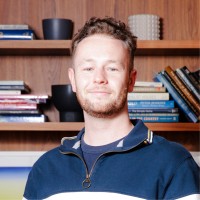 A portrait photo of Louis Docherty standing indoors in front of a bookshelf, wearing a casual top. The background features neatly arranged books, creating a warm and professional setting.
